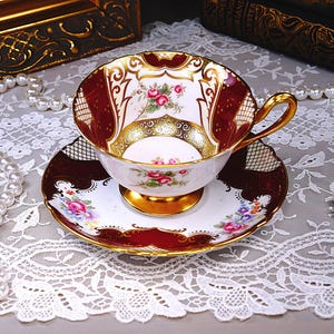 Può includere: Un set vintage di tazza e piattino da tè. La tazza presenta un manico e un bordo dorati, con un design bordeaux e oro. Il piattino ha un bordo bordeaux e oro abbinato, con un motivo floreale al centro. Il set è posto su una tovaglia di pizzo bianco.