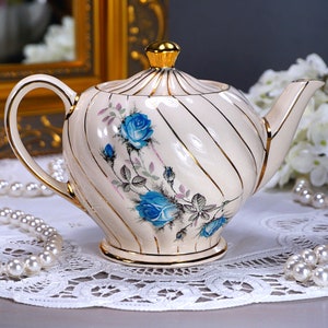 May include: A white teapot with gold trim and blue rose floral design. The teapot has a gold lid and a curved spout.