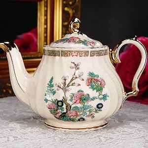 May include: A white teapot with a floral design featuring pink and green flowers and gold accents. The teapot has a gold handle and a gold lid.