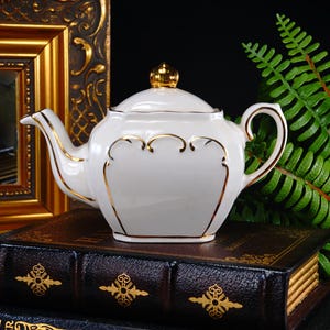 May include: A white porcelain teapot with gold trim and a gold finial on the lid. The teapot is sitting on a stack of antique books with gold accents. A gold framed mirror and green fern are in the background.
