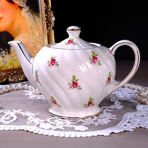May include: A white porcelain teapot with a lid, decorated with pink rose patterns and gold edging. The teapot is on a white lace doily, with a pearl necklace and a framed portrait in the background. A classic tea set, perfect for serving tea.