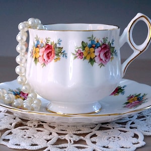 May include: A white teacup and saucer with a floral pattern in shades of pink, yellow, and blue. A pearl necklace is draped over the cup and saucer.