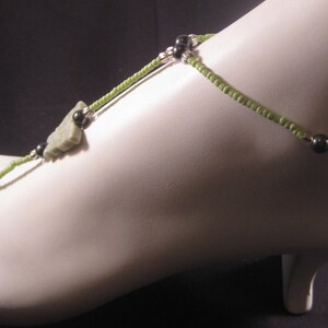 May include: A green beaded ankle bracelet with a silver chain and a green stone charm. The bracelet is worn on a mannequin's foot.