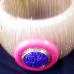 May include: A wooden bangle bracelet with a pink and purple button. The button has a gold rim and a floral design.
