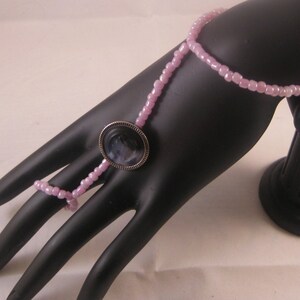 May include: A pink beaded finger ring with a round, silver-toned, black cameo-style pendant.