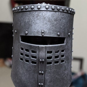 May include: A silver metal helmet with a visor and a grid pattern on the front. The helmet has a textured surface and is decorated with rivets.