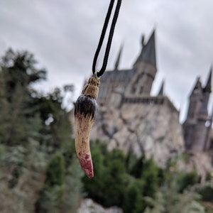 Puede incluir: Un colgante en forma de diente marrón y negro con una mancha roja en la punta. El colgante cuelga frente a un fondo borroso de un edificio de piedra.