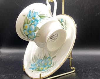 May include: A white teacup and saucer set featuring a floral pattern of blue flowers and green leaves. The set is displayed on a gold-coloured metal stand. The teacup has a curved handle and gold trim. The saucer also has a gold trim.