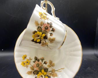 May include: White teacup and saucer set with a floral design featuring yellow and peach roses and gold trim. The teacup is resting on a gold-colored stand. The saucer has a scalloped edge.