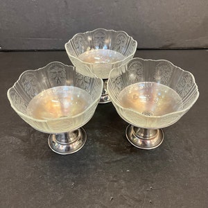 Peut inclure: Trois coupes &agrave; dessert en verre transparent avec des bases argent&eacute;es. Les coupes ont un bord festonn&eacute; et un motif grav&eacute; d&eacute;licat.