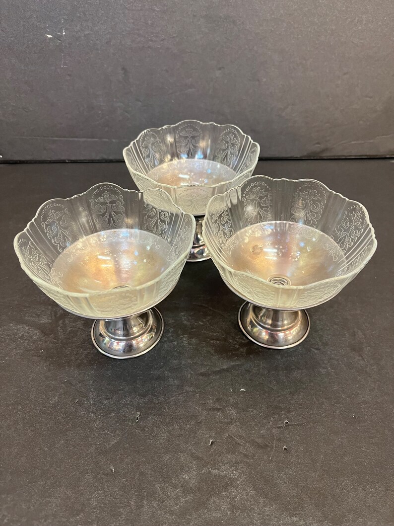 Peut inclure: Trois coupes &agrave; dessert en verre transparent avec des bases argent&eacute;es. Les coupes ont un bord festonn&eacute; et un motif grav&eacute; d&eacute;licat.
