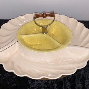 May include: A three-tiered ceramic serving dish shaped like a seashell. The top tier is yellow and divided into three sections. The bottom two tiers are a pale pink colour. A wooden handle is attached to the top tier.