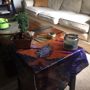 May include: A wooden coffee table with a dark brown tabletop and black legs. The table is covered with a colourful tablecloth featuring a purple, orange and blue design. A small potted bonsai tree sits on the table, along with a glass jar with a teal candle inside.