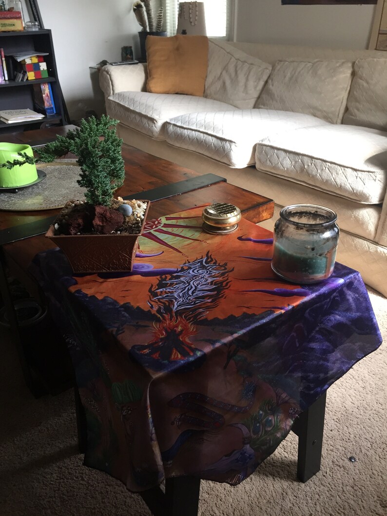 May include: A wooden coffee table with a dark brown tabletop and black legs. The table is covered with a colourful tablecloth featuring a purple, orange and blue design. A small potted bonsai tree sits on the table, along with a glass jar with a teal candle inside.