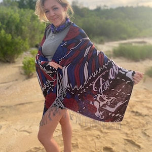 May include: A woman wearing a gray shirt and a colorful shawl with fringe. The shawl has a floral pattern and the text "Faerie" on it. She is standing on a sandy beach.