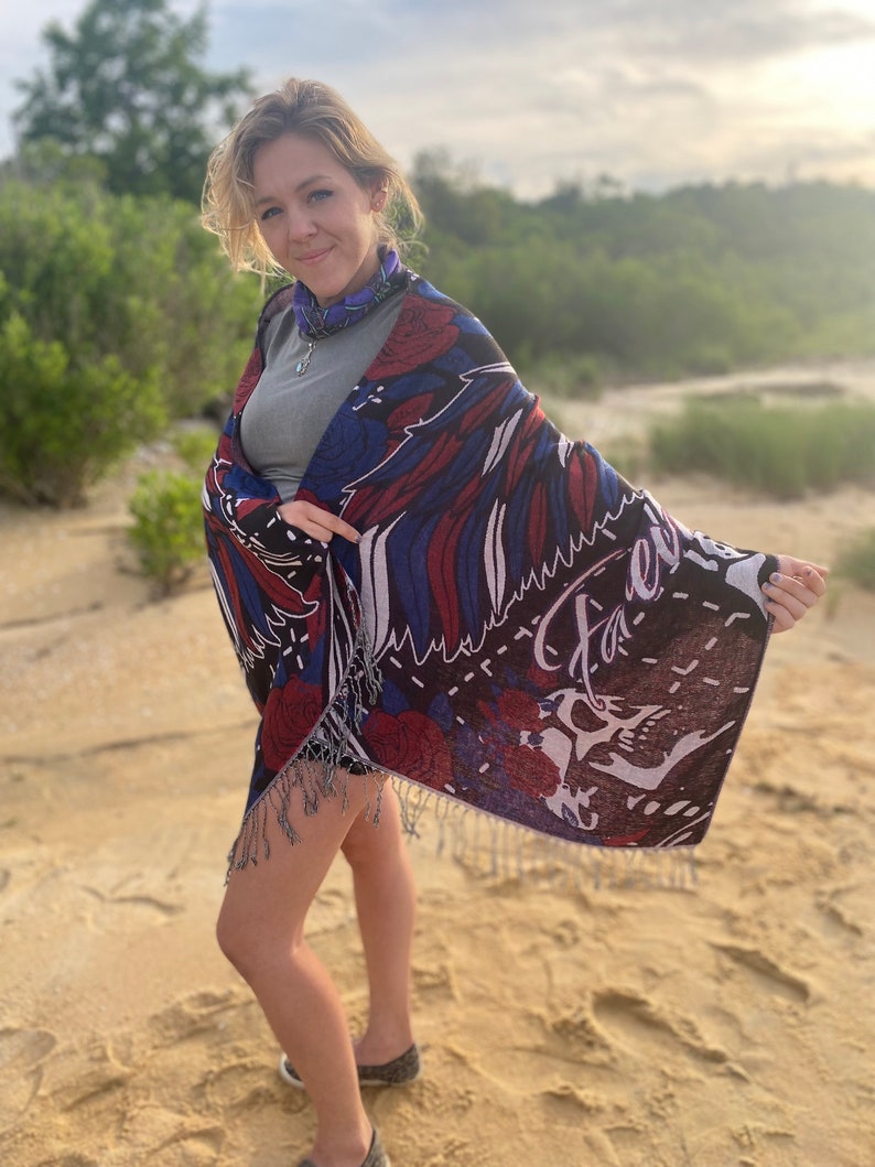 May include: A woman wearing a gray shirt and a colorful shawl with fringe. The shawl has a floral pattern and the text "Faerie" on it. She is standing on a sandy beach.