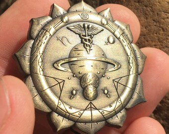 May include: A gold-toned metal pin with a detailed design featuring a stylized caduceus, a planet with rings, and other astrological symbols. The pin is shaped like a flower with multiple petals.