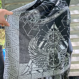 May include: Black and white geometric patterned scarf with a large, detailed design of a winged scarab beetle and a flower of life symbol. The scarf has fringe on the edges.
