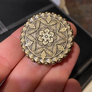 May include: A gold-toned metal medallion with a detailed geometric design featuring a star of David in the center. The design includes various shapes, including triangles, squares, and circles.