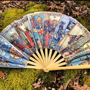 May include: A hand fan made of bamboo with a colorful design featuring the Major Arcana cards from a tarot deck. The cards include The Magician, The Empress, The Lovers, Judgement, Death, The Fool, The Sun, and The World.
