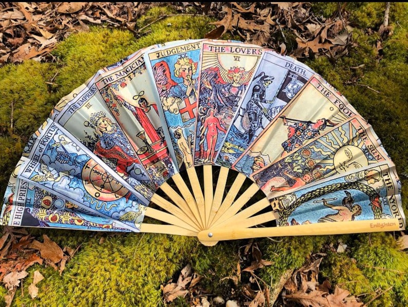 May include: A hand fan made of bamboo with a colorful design featuring the Major Arcana cards from a tarot deck. The cards include The Magician, The Empress, The Lovers, Judgement, Death, The Fool, The Sun, and The World.