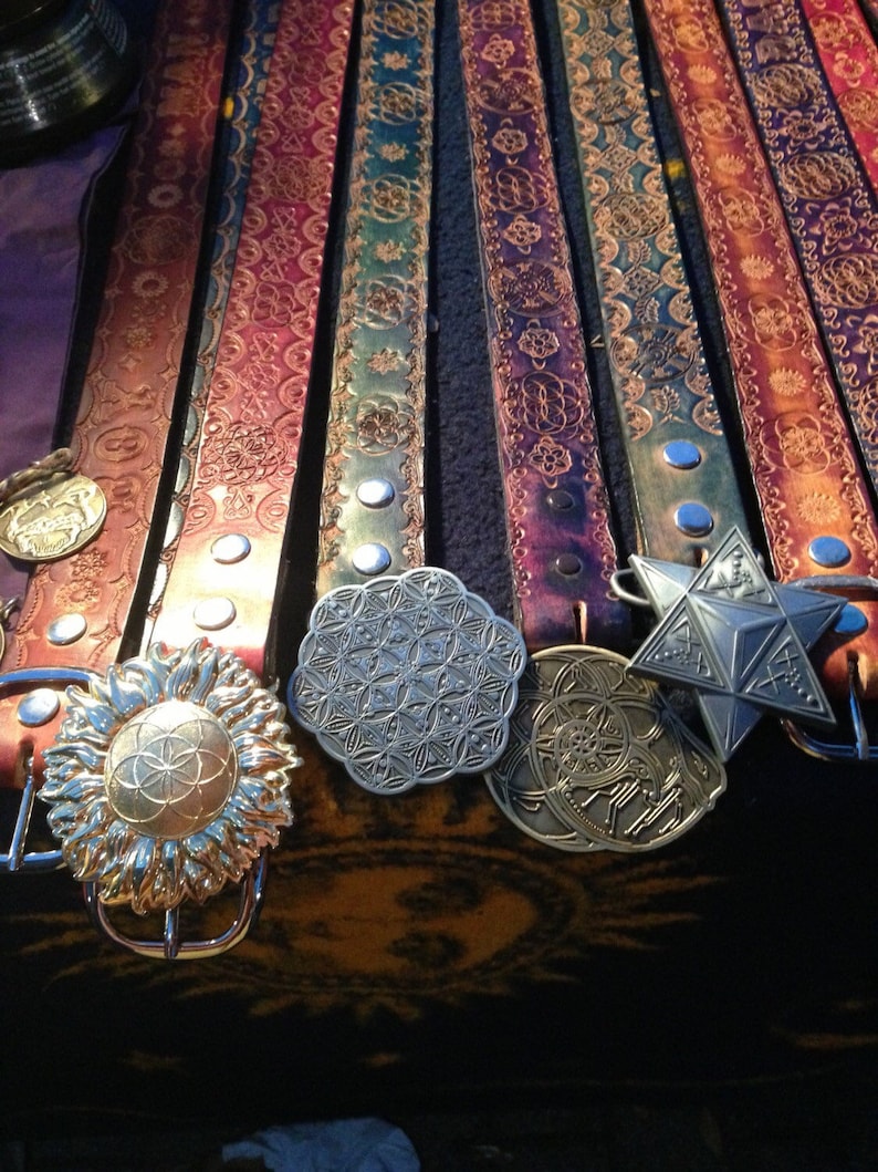 May include: A collection of eight leather belts with intricate designs and metal buckles. The belts are arranged in a row, with the buckles facing the viewer. The buckles are all different shapes and sizes, and some have a flower or geometric pattern.