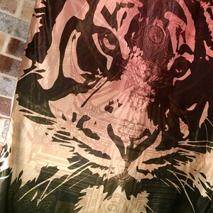 May include: A fabric with a tiger face design. The tiger's face is depicted in black and brown against a light pink and beige background. The design includes intricate patterns and details, creating a striking visual.