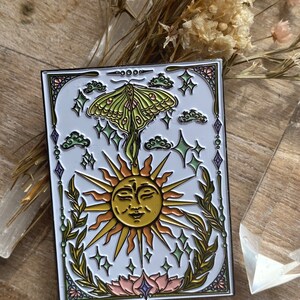 May include: A green and gold enamel pin featuring a stylized sun with a face, a green moth with pink accents, and a floral design. The pin is surrounded by a black border and has a white background.
