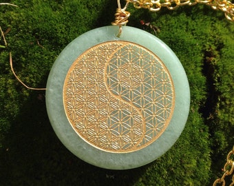 May include: A green stone pendant featuring a gold-coloured yin yang symbol and a flower of life design. The pendant is attached to a gold chain.