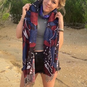 May include: A woman wearing a black and blue floral patterned scarf with fringe. The scarf has red, white, and blue accents. She is wearing a gray tank top and denim shorts.