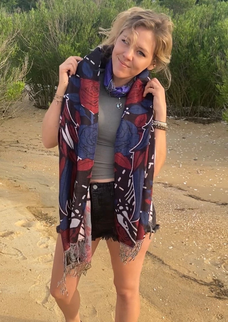 May include: A woman wearing a black and blue floral patterned scarf with fringe. The scarf has red, white, and blue accents. She is wearing a gray tank top and denim shorts.