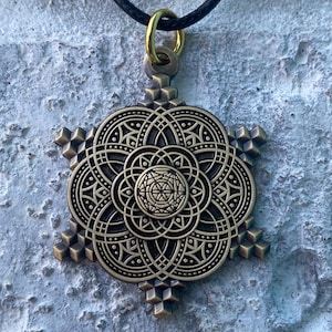 May include: A bronze-colored pendant with an intricate, symmetrical design, suspended from a black cord. The pendant features a central circular pattern surrounded by detailed geometric shapes and six outward-pointing, cube-like structures. A gold-colored ring connects the pendant to the cord.