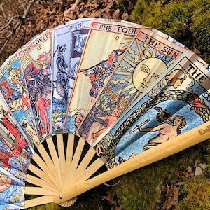May include: A hand fan made of bamboo with a design of tarot cards. The cards are in a fan shape and show the following cards: Judgement, The Lovers, Death, The Fool, The Sun, and The World. The fan is made of wood and has a light brown color.