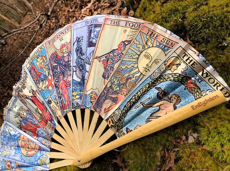 May include: A hand fan made of bamboo with a design of tarot cards. The cards are in a fan shape and show the following cards: Judgement, The Lovers, Death, The Fool, The Sun, and The World. The fan is made of wood and has a light brown color.