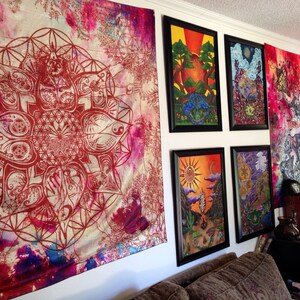 May include: A room featuring wall art and tapestries. A large tapestry with a red geometric design hangs on the left. Several framed art pieces with colorful landscapes are displayed, along with a vibrant tapestry on the right.
