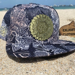 May include: A black and white patterned baseball cap with a gold geometric design on the front. The cap has a curved brim and a flat top. The text "Enlighten Clothing Co." is visible on a wooden sign in the background.