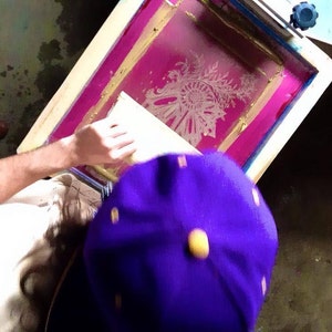 May include: A person is using a screen printing frame with a pink background and a white design. The frame is made of wood and has a metal handle. The person is wearing a purple hat.