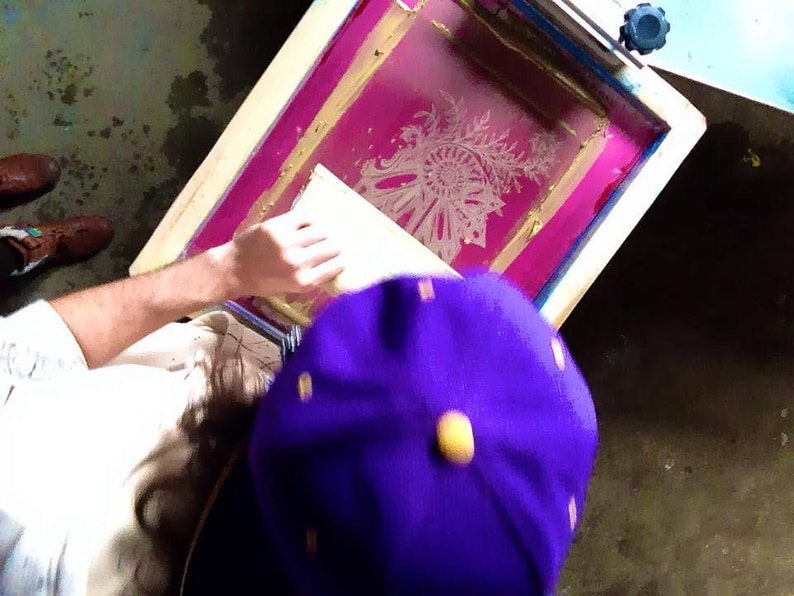 May include: A person is using a screen printing frame with a pink background and a white design. The frame is made of wood and has a metal handle. The person is wearing a purple hat.
