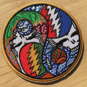 May include: A circular embroidered patch with a colorful design featuring a skull, lightning bolts, and a swirling pattern. The patch is surrounded by a gold border.