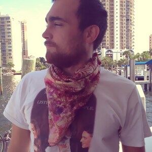 May include: A man wearing a white t-shirt with a floral print scarf and the text "Listen to the People." The shirt also features a graphic of two women with a bottle of beer.