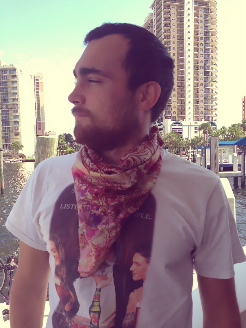 May include: A man wearing a white t-shirt with a floral print scarf and the text "Listen to the People." The shirt also features a graphic of two women with a bottle of beer.