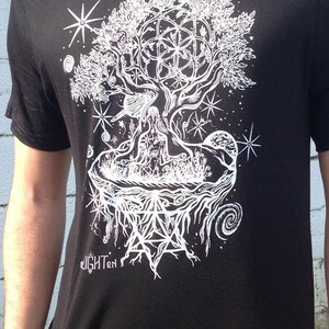 Pode incluir: Camiseta preta com um gr&aacute;fico branco de uma &aacute;rvore com detalhes intrincados, incluindo um design de flor da vida, estrelas e um padr&atilde;o geom&eacute;trico. O texto "Enlighten" &eacute; vis&iacute;vel na parte inferior do design.
