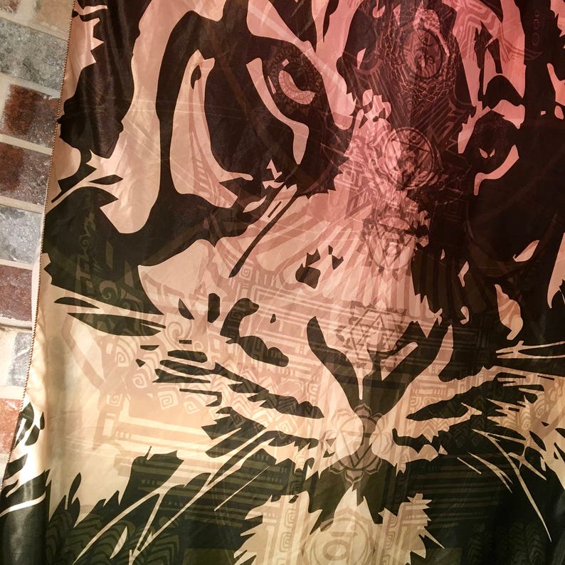 May include: A fabric with a tiger face design. The tiger's face is depicted in shades of black and brown, with a light pink and beige background. The fabric has a detailed, artistic pattern, creating a striking visual effect.