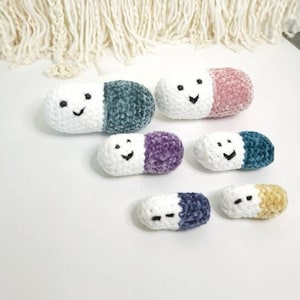 May include: A set of six crocheted pill-shaped plushies in various colors, including white, pink, purple, and blue. Each plushie has a smiling face.