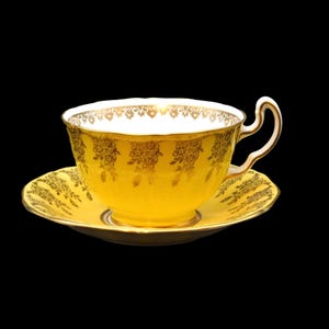 May include: A yellow teacup and saucer set with gold floral designs. The teacup has a curved handle and a white interior rim with gold trim. The saucer also has gold trim and floral patterns. The set is against a black background.