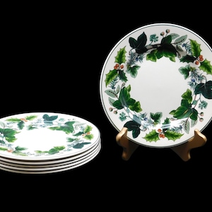 May include: Set of white ceramic plates with a green and red holly leaf design. One plate is displayed on a gold stand, while a stack of four plates is visible on the left. The plates have a black trim.