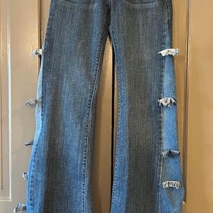 May include: Blue denim flared jeans with light blue side panels and decorative fabric bows. The jeans have a classic five-pocket design and are hanging on a wooden hanger. The bottom hems are frayed.