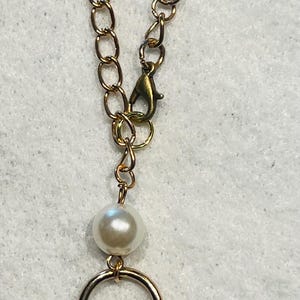 May include: Gold-toned chain with a lobster clasp, a large pearl, and a gold ring. A smaller pearl hangs below the ring. The jewelry is set against a white background.