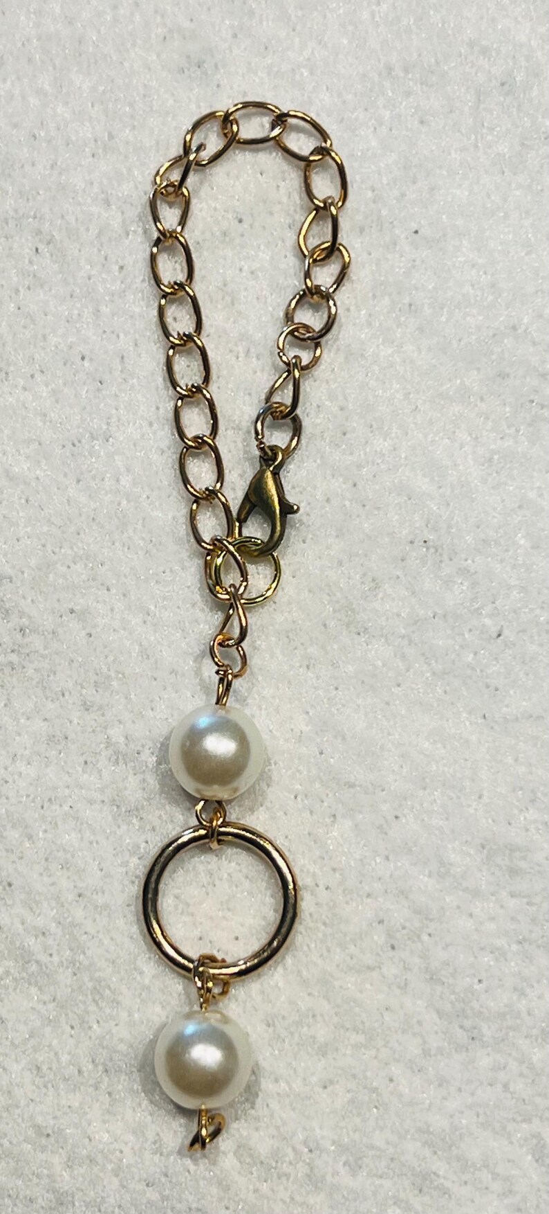 May include: Gold-toned chain with a lobster clasp, a large pearl, and a gold ring. A smaller pearl hangs below the ring. The jewelry is set against a white background.