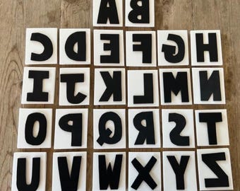 May include: Black uppercase letters on white square tiles arranged on a wooden surface. The letters are in a bold, sans-serif font. The tiles are evenly spaced, creating a grid-like pattern. The letters are used for educational purposes.